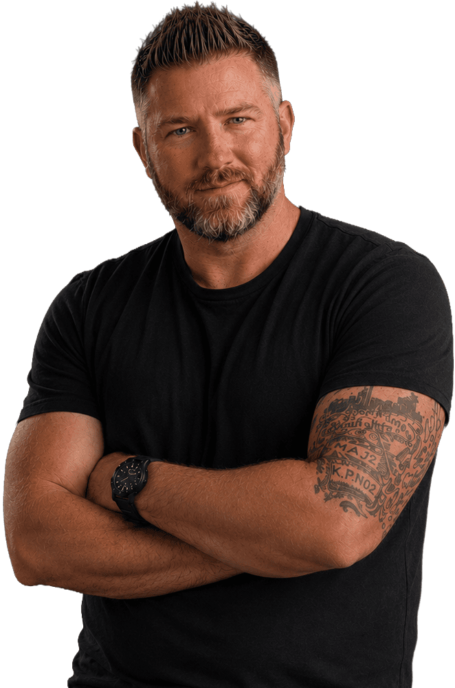 A man with short hair and a beard stands with arms crossed, wearing a black t-shirt and watch, showing a detailed tattoo on his arm. He has a serious expression and looks directly at the camera.