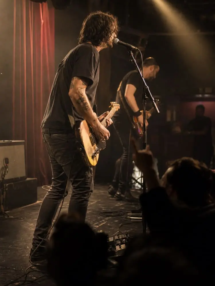 Two musicians play electric guitars and sing on a dimly lit stage. One stands at the microphone in the foreground, while an audience member raises a hand in front of the stage.