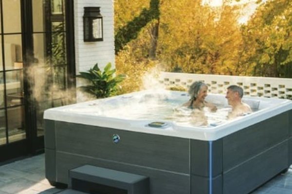 Two people relax in a steaming outdoor hot tub on a patio near a house, surrounded by greenery and warm sunlight.